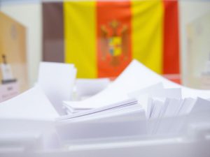 Urnă de vot, buletine de vot, steag românesc, oameni votând.