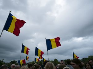 Oameni adunați, steaguri românești fluturând, nori întunecați.