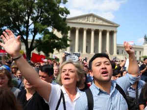 Oamenii protestând furioși, clădiri guvernamentale în fundal.